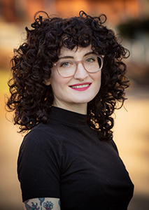 Headshot of Jill Forgash a young woman with curly brown hair and glasses wearing a fitted black turtle neck with a soft open mouth smile.