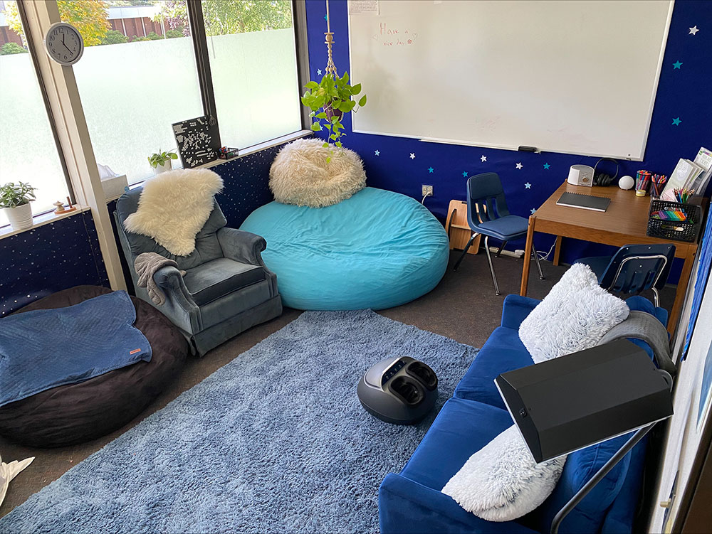 Open and inviting sensory room featuring bean bags, a couch, and chairs. A desk adorned with coloring utensils, a journal, headphones, and more. The space is enhanced with decorative plants, a painted wall embellished with star stickers and a whiteboard.