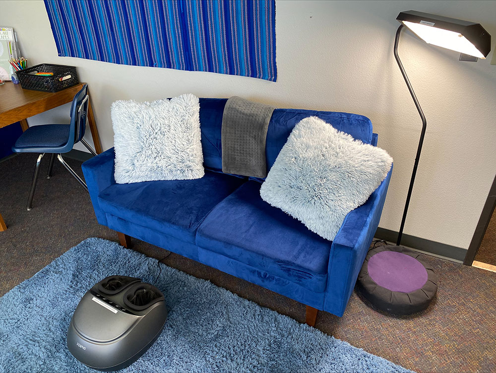Comfortable seating arrangement featuring a couch adorned with pillows and a foot massager at one end. Next to it is a side lamp.