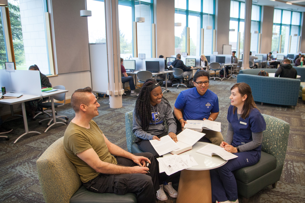 Four students talking, smiling while sitting around a desk with text books and papers in Cannell Library