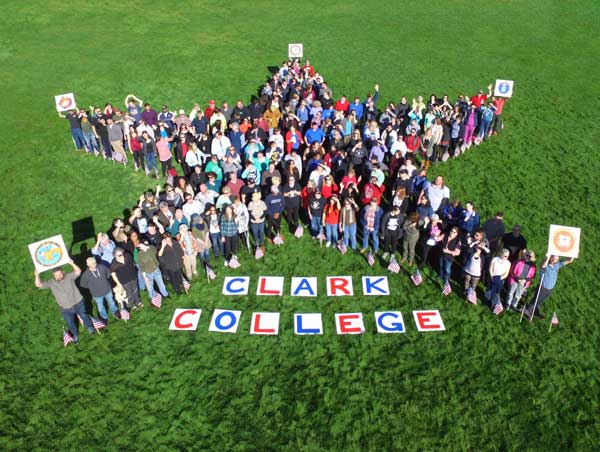 A birds eye view of a group of people huddled together, forming a star, with large paper letters that spell 'Clark College', and smiling up as they get their picture taken on Veterans Day
