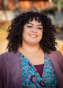 Katia Quintero headshot. Katia is wearing a pink blazer and patterned shirt smiling with teeth. Her hair is curly and shoulder length. 