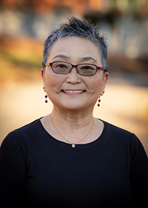 Lana Strickland Headshot features a middle aged woman with short hair and glasses, smiling in an inviting way with direct eye contact wearing a crew neck black shirt.