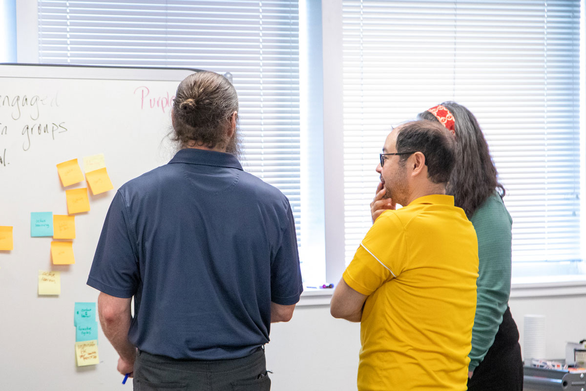 Three people standing together, looking pensive as they examine a whiteboard covered with sticky notes on it.