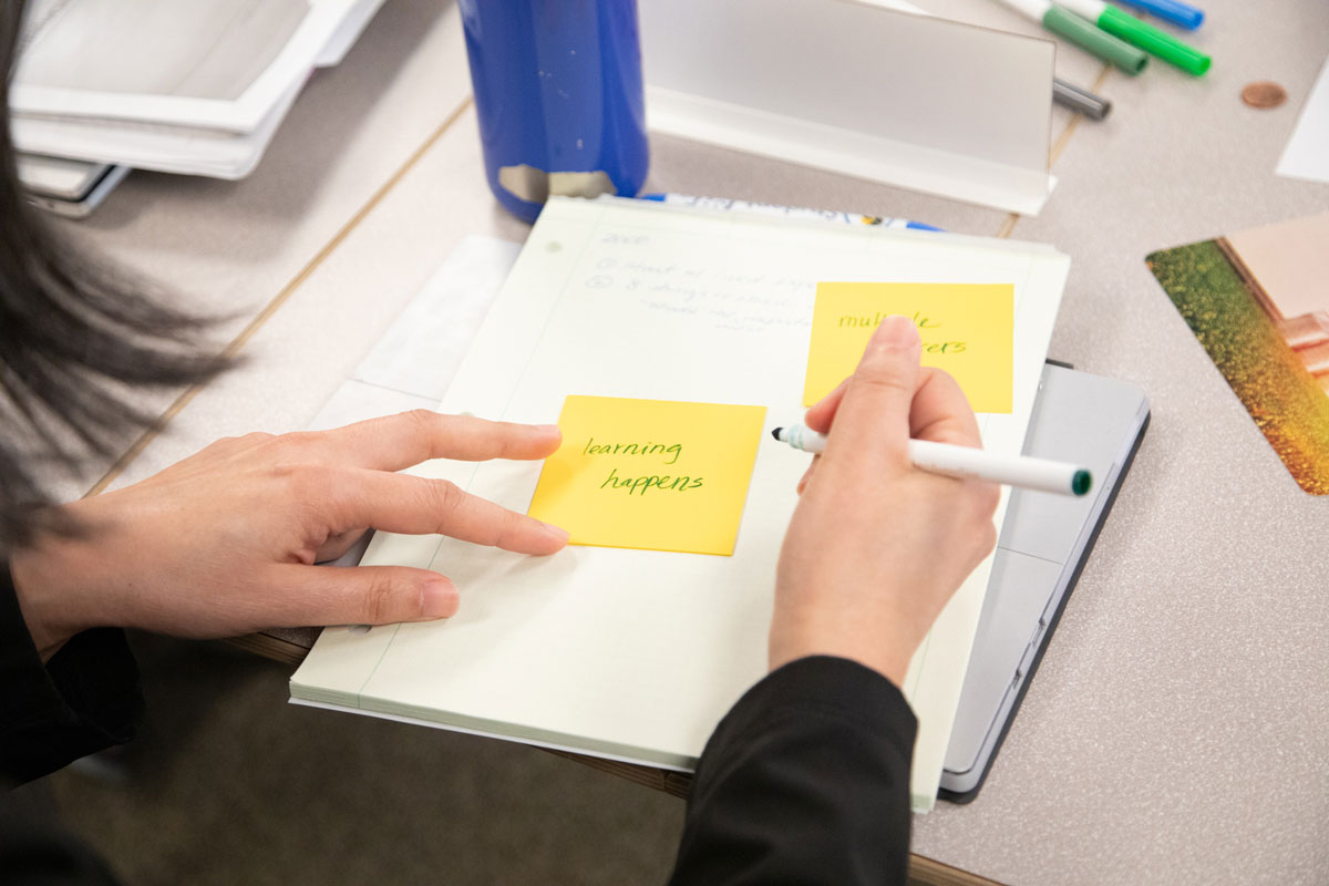 A person at a desk with a notebook, holding a marker over a sticky note with the words 'Learning happens' written on it.
