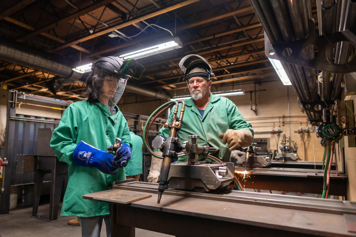 Two individuals wearing protective gear, including visor helmets and heavy-duty gloves, operate a large welding machine indoors.
