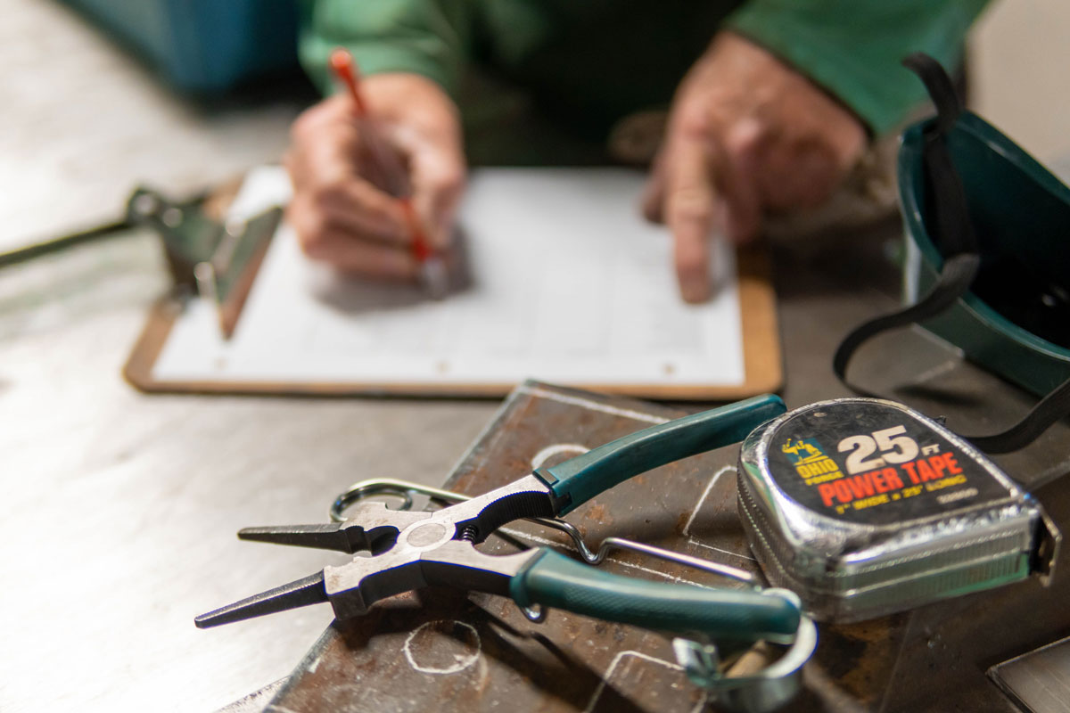 A pair of pliers and a measuring tape in focus, with a blurred foreground showing someone writing on a clipboard.