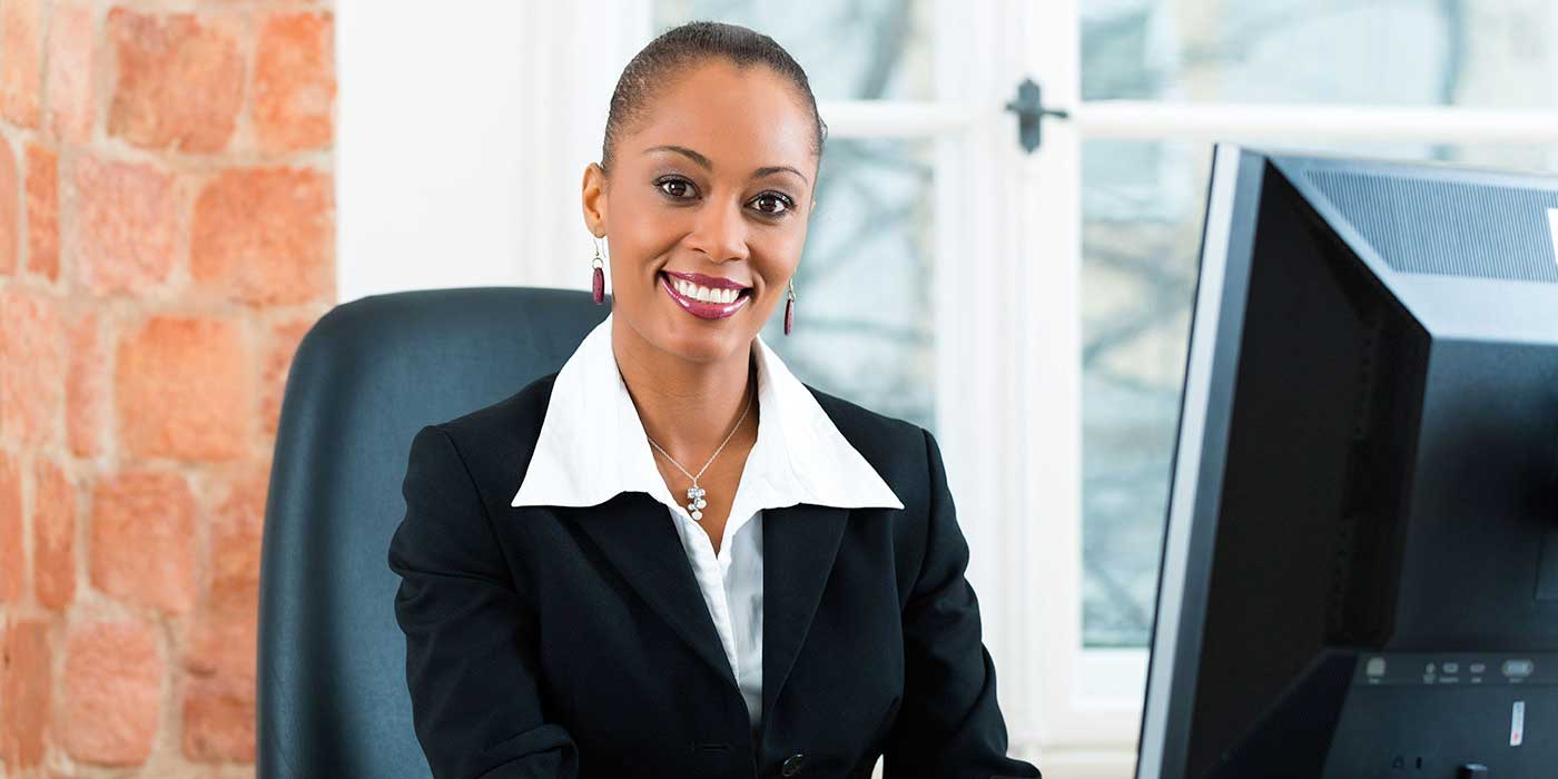 A woman in formal attire sitting at a desk, smiling at the camera while working on a computer.