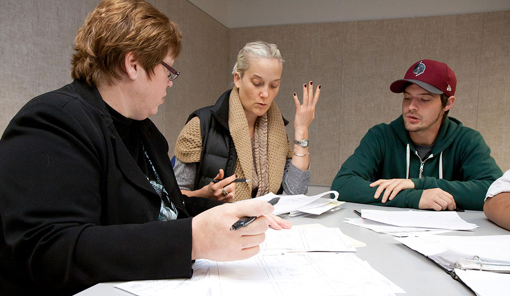 three people at a table with papers and pens on the table and one person talking