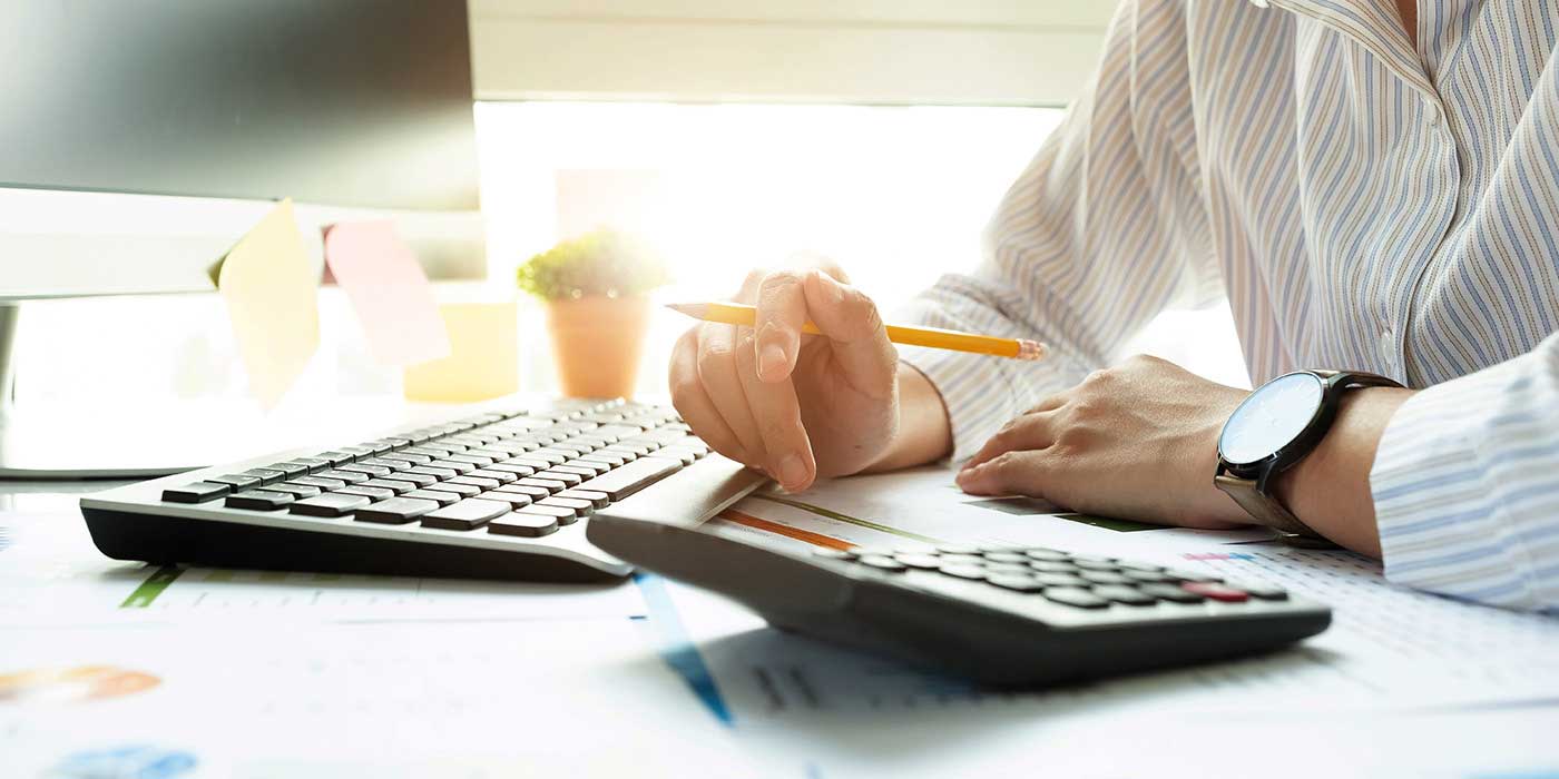 Close-up of hands holding a pencil on a desk, with a computer, keyboard, and calculator nearby.
