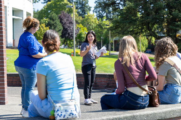 A woman holding papers speaks to four people, three of which are seated on a bench against a wall.