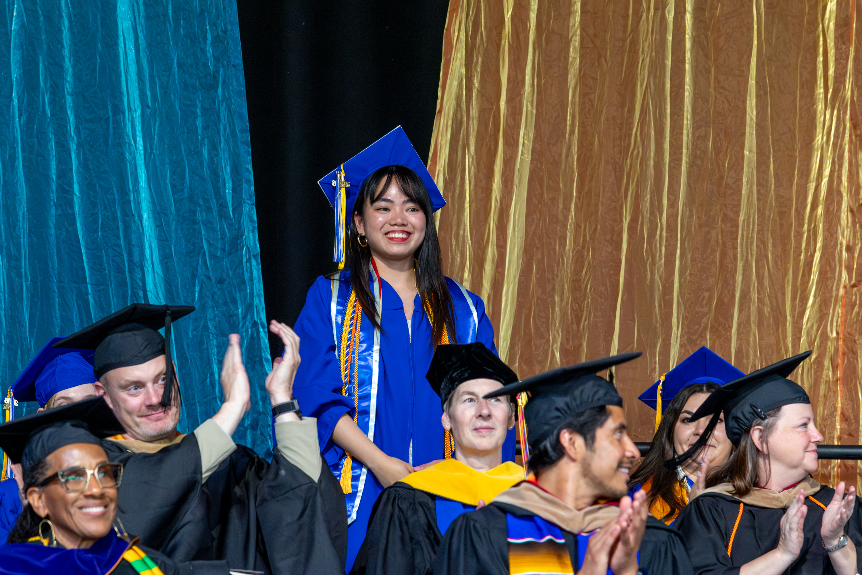Photo of a student in regalia standing among clapping faculty members and students.