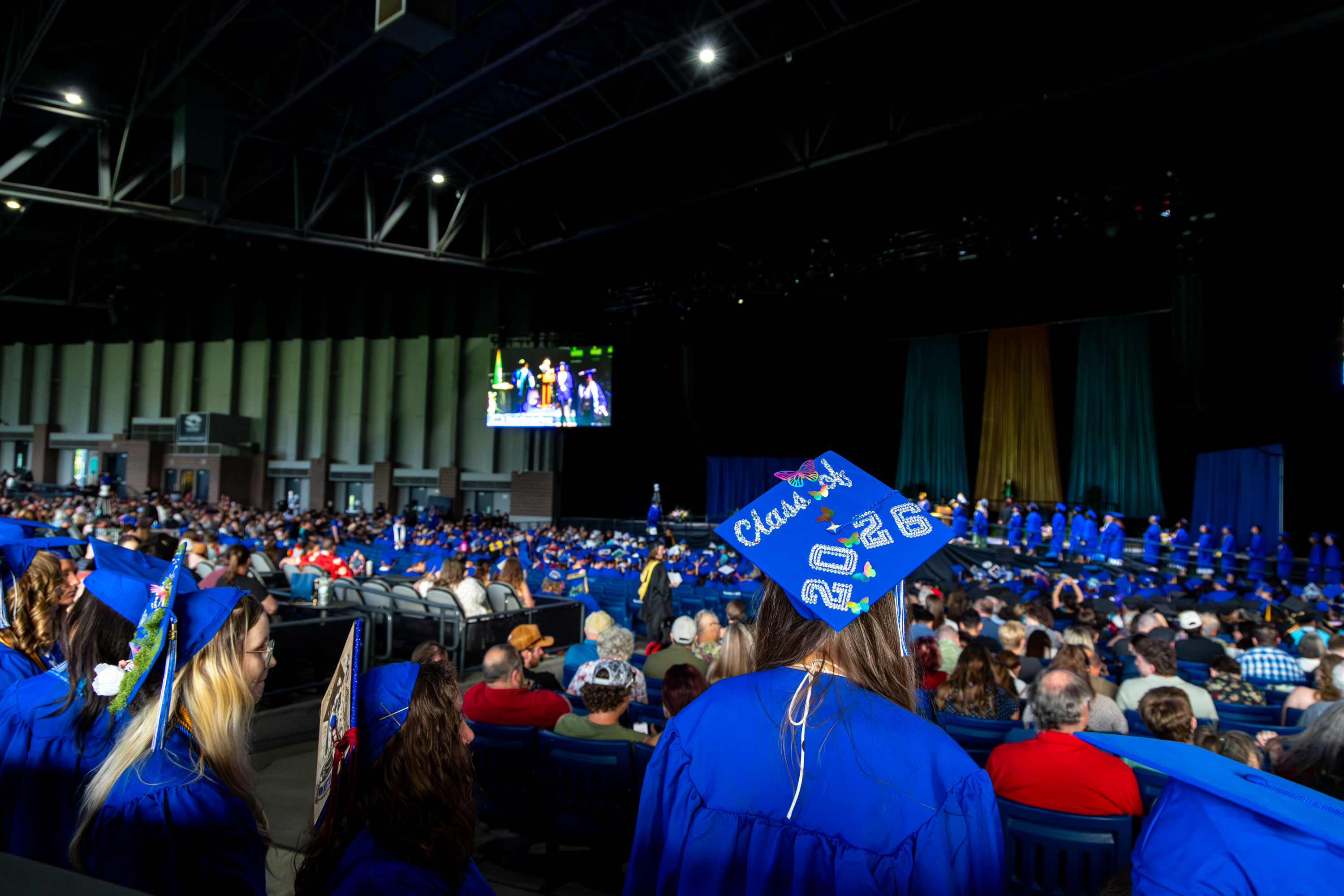 Photo of a crowd of graduates in blue regalia, with a decorated graduation cap front and center that says "Class of 2026"