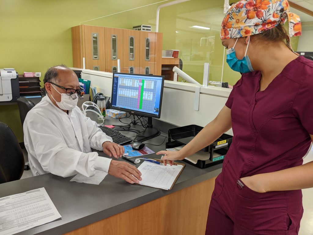 Dentist and dental hygiene student, both wearing masks, look at clipboard together.