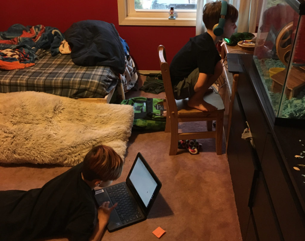 two boys looking at computer screens for online school in a bedroom