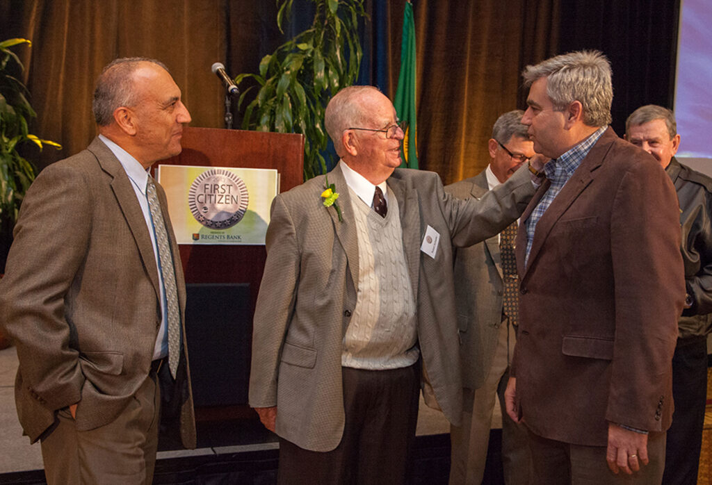 A person placing a hand on Bob Knight's shoulder. A sign on a podium behind them "2013 First Citizen".