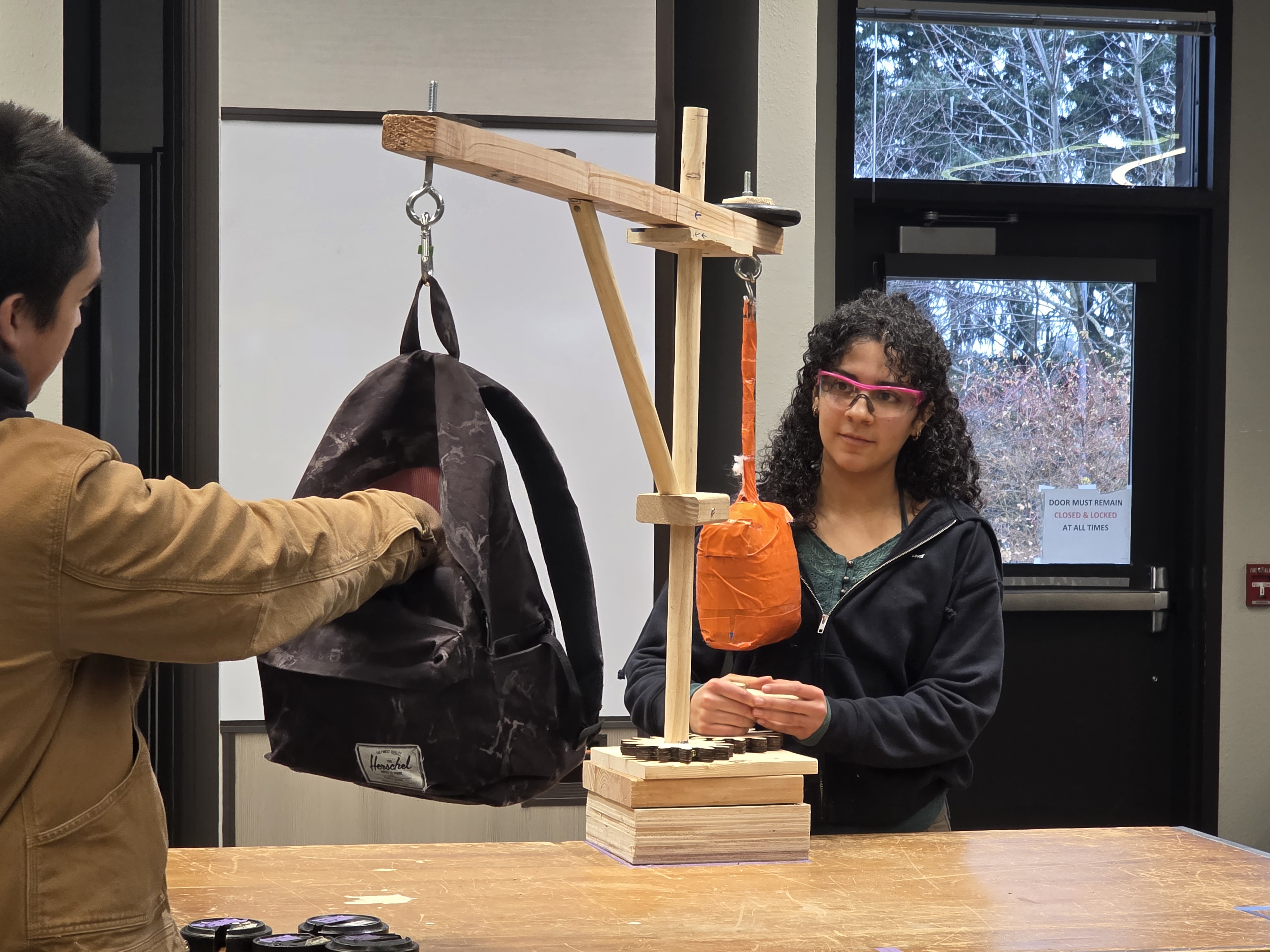 Student wearing safety glasses operating crane while another student loads weight into the attached bag 