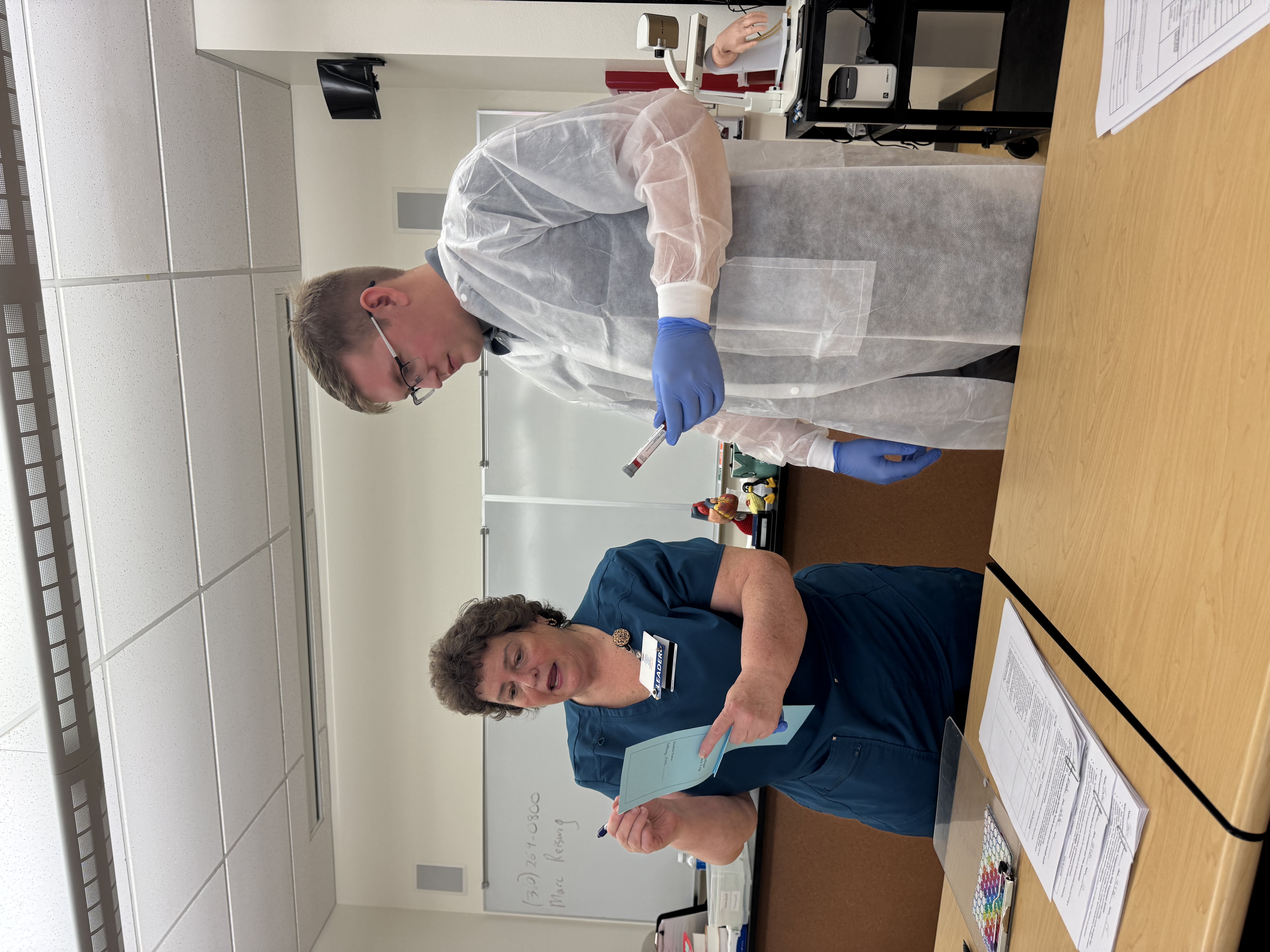 Phlebotomy faculty member references live blood draw log as state trooper watches, holding vial of blood