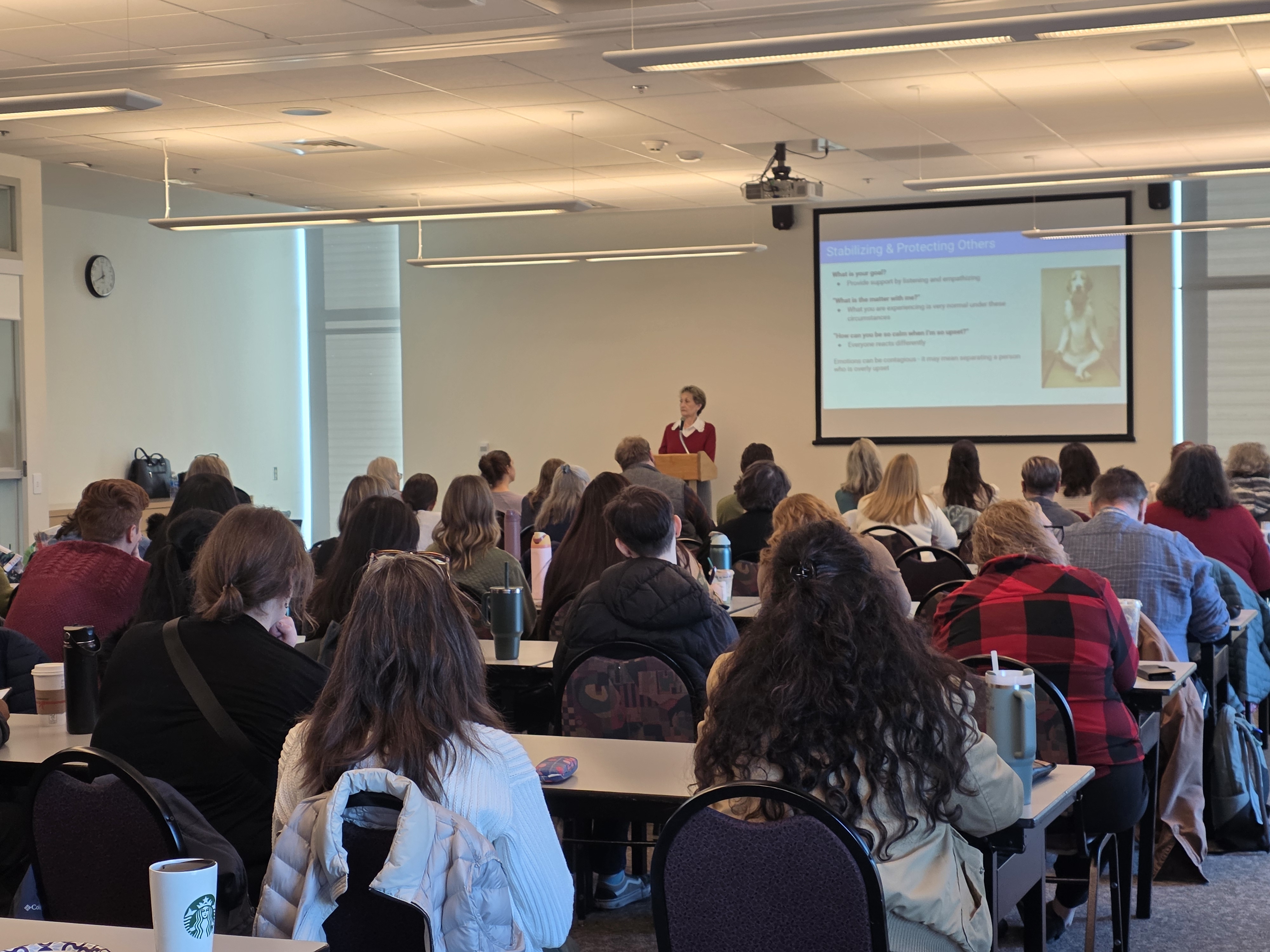Classroom of people looking at a speaker at a podium in front of a projection screen