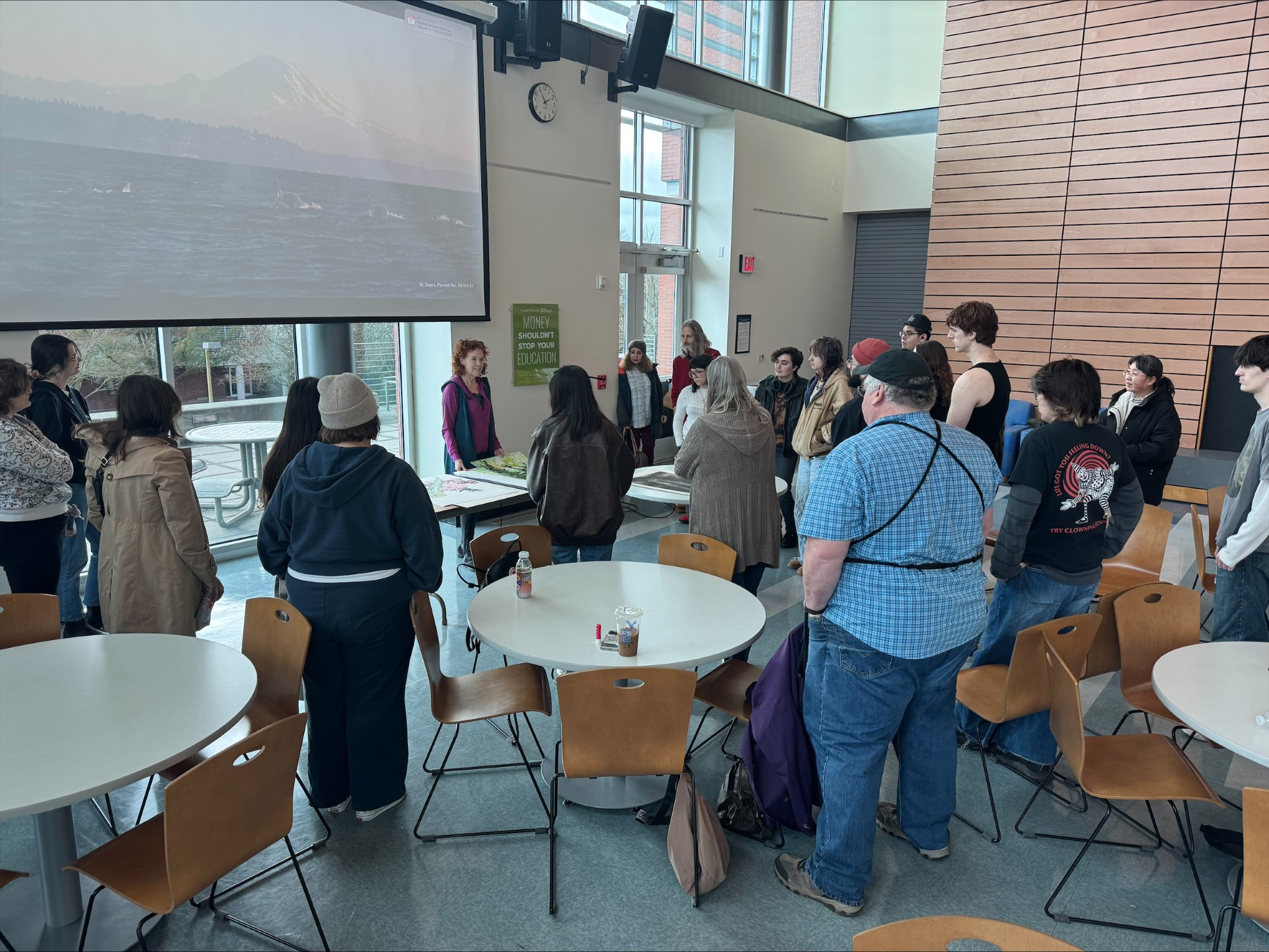 People gathered around an individual at the front of a room, who is motioning to artwork on the table in front of her.