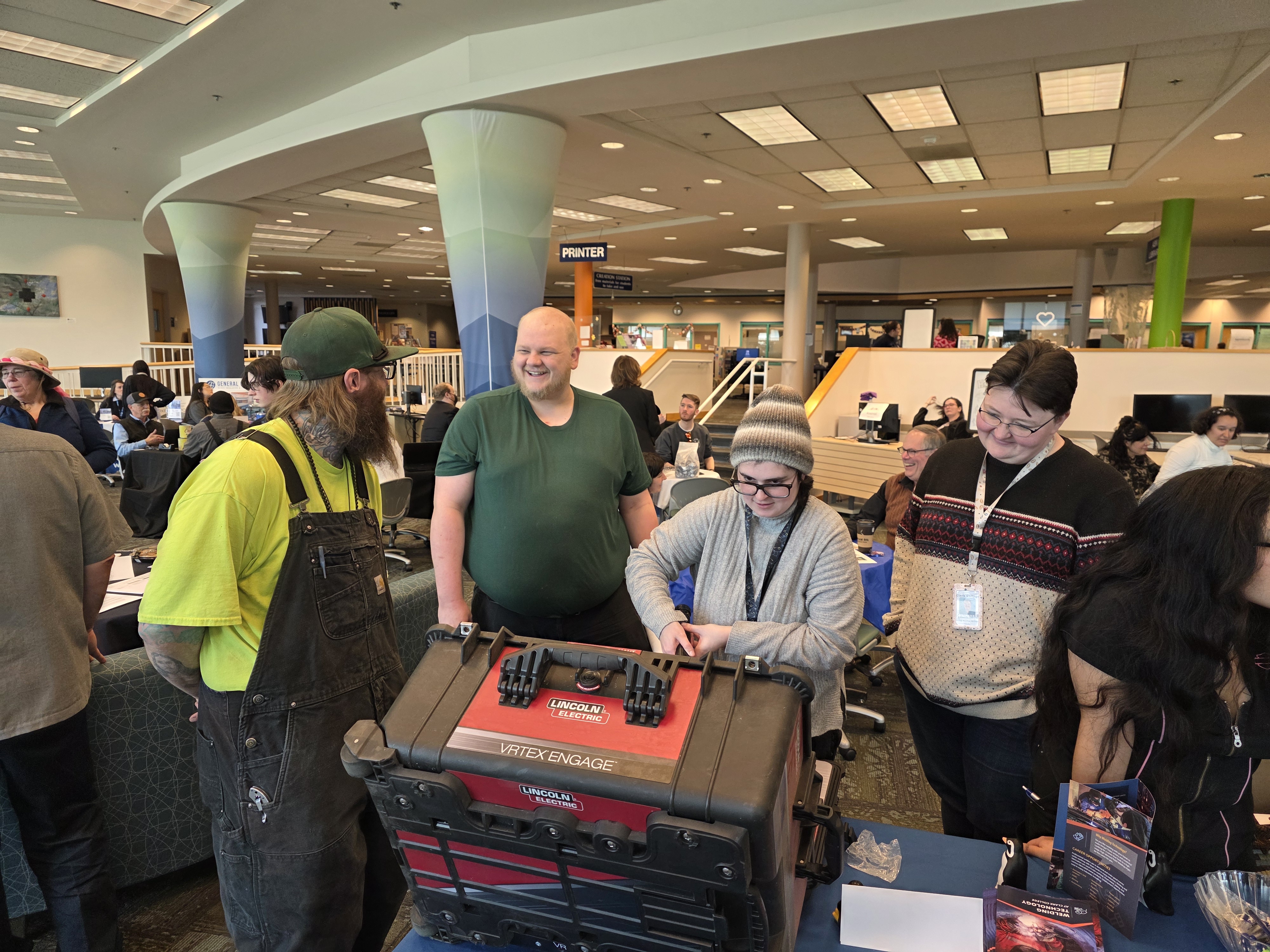A group of people stand in front of a welding simulator on a table as one of them is holding the welder.