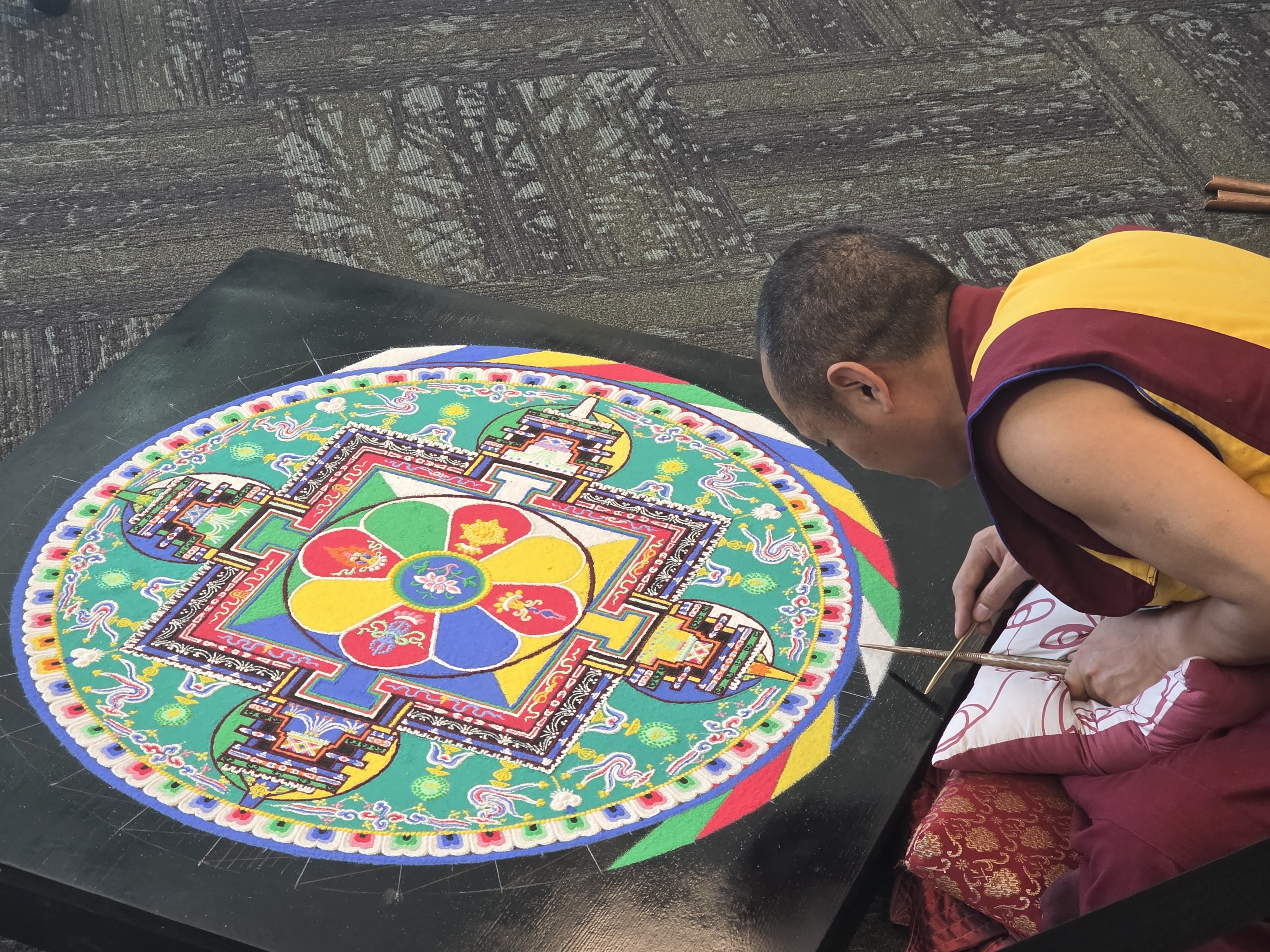 Monk in maroon and yellow robe sitting on the floor, leaning over a black block as he adds sand to a mandala