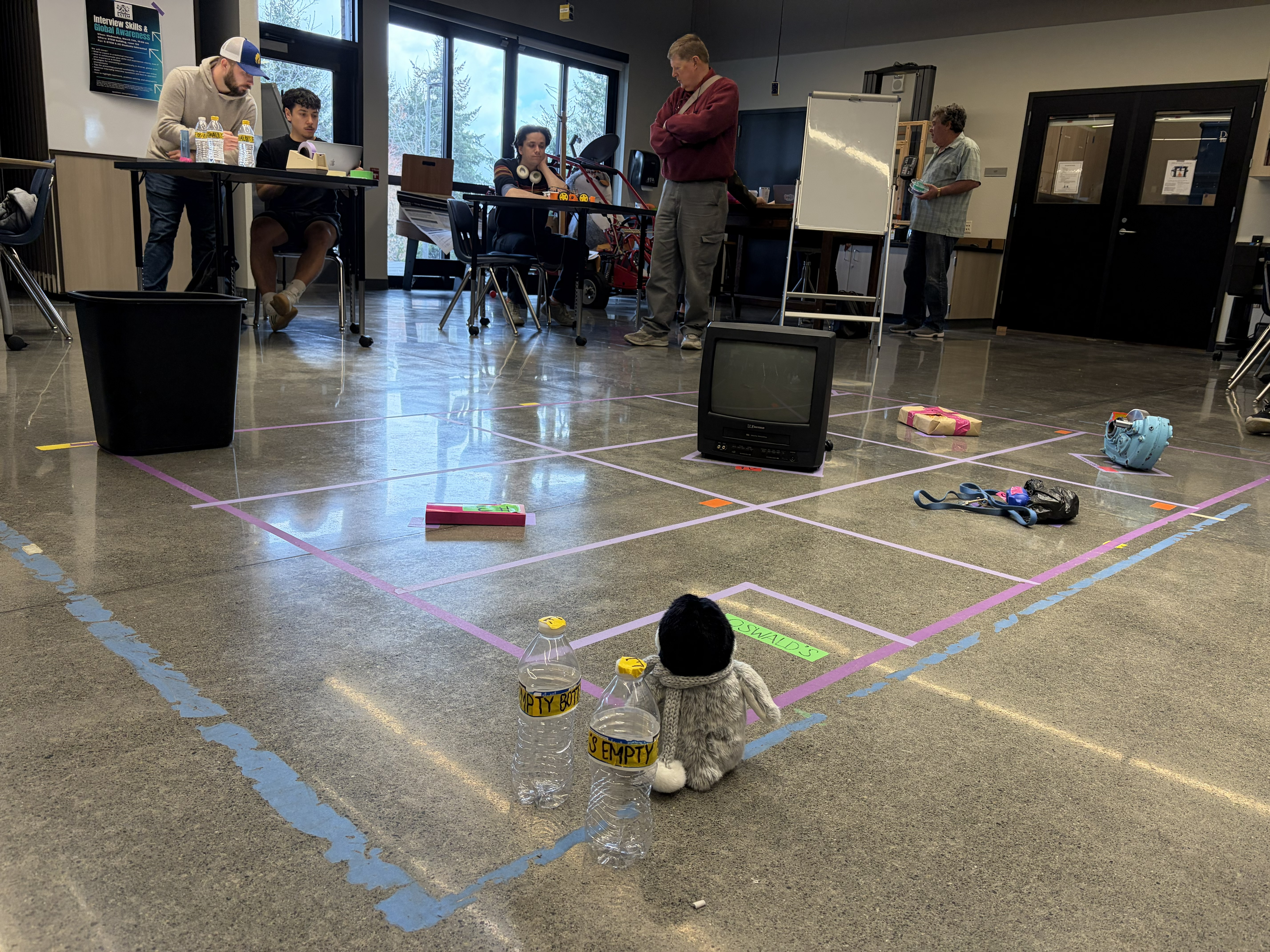 Giant square outlined on floor with colorful tape and small obstacles scattered throughout the space, including a small TV. Stuffed Oswald sits on one side of the square.