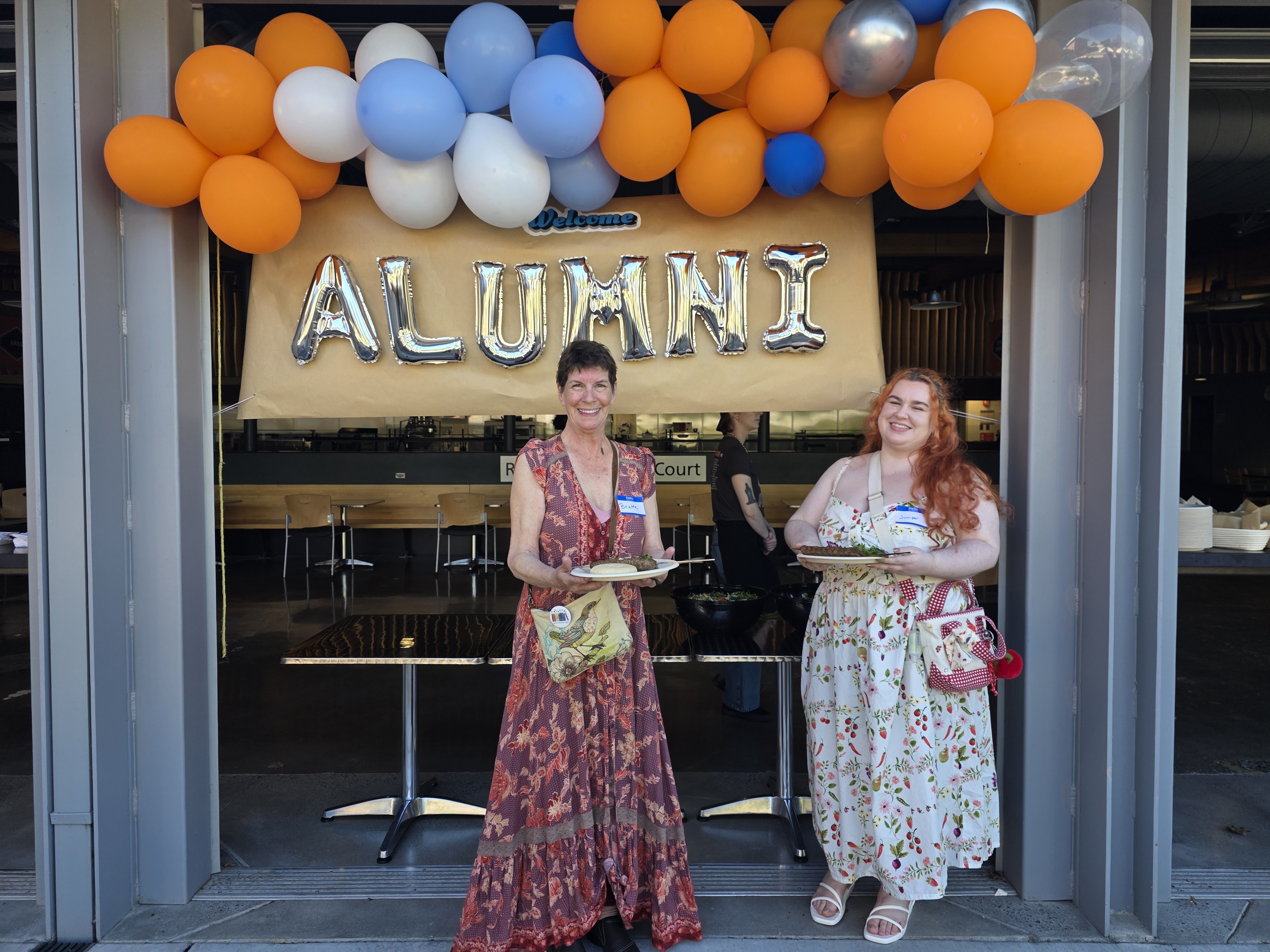 Two culinary alum stand under a sign that says "alum" with blue and orange balloons. Both are holding plates of food.