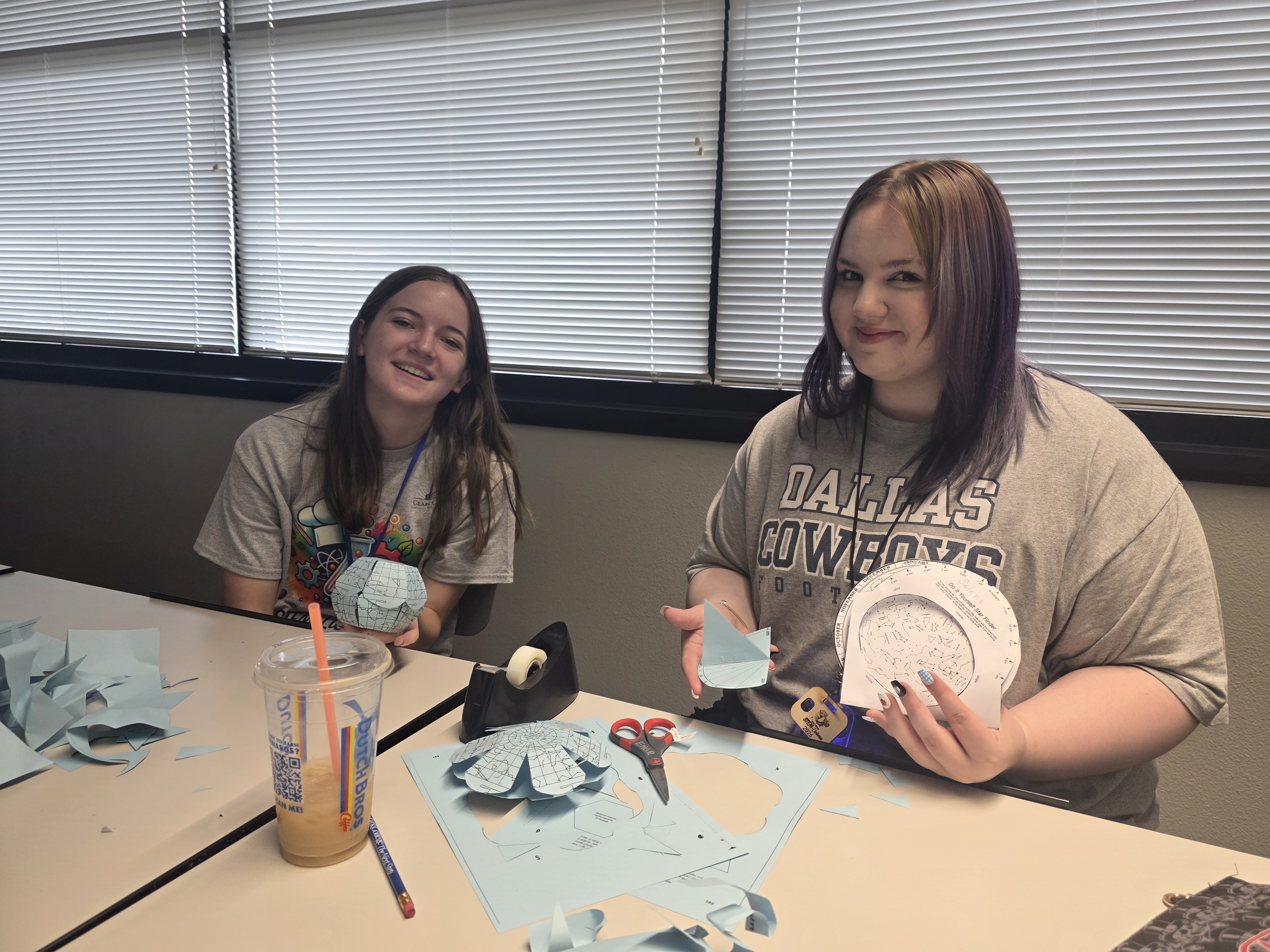 Two students sit at a table, holding paper astronomy projects.