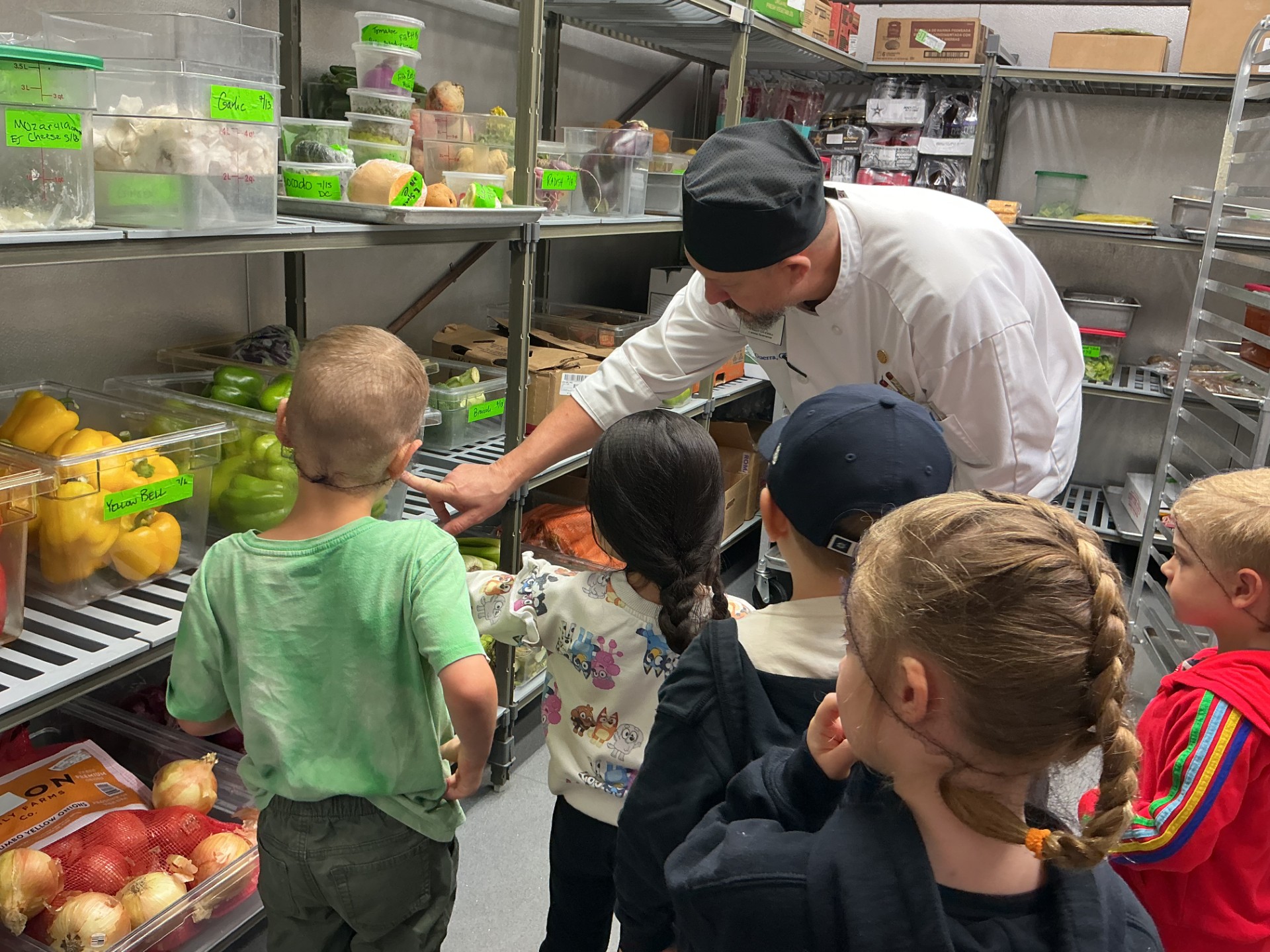Chef Aaron points to a container of fruit on a shelf in the MCI kitchen refridgerator, with a group of children looking on.