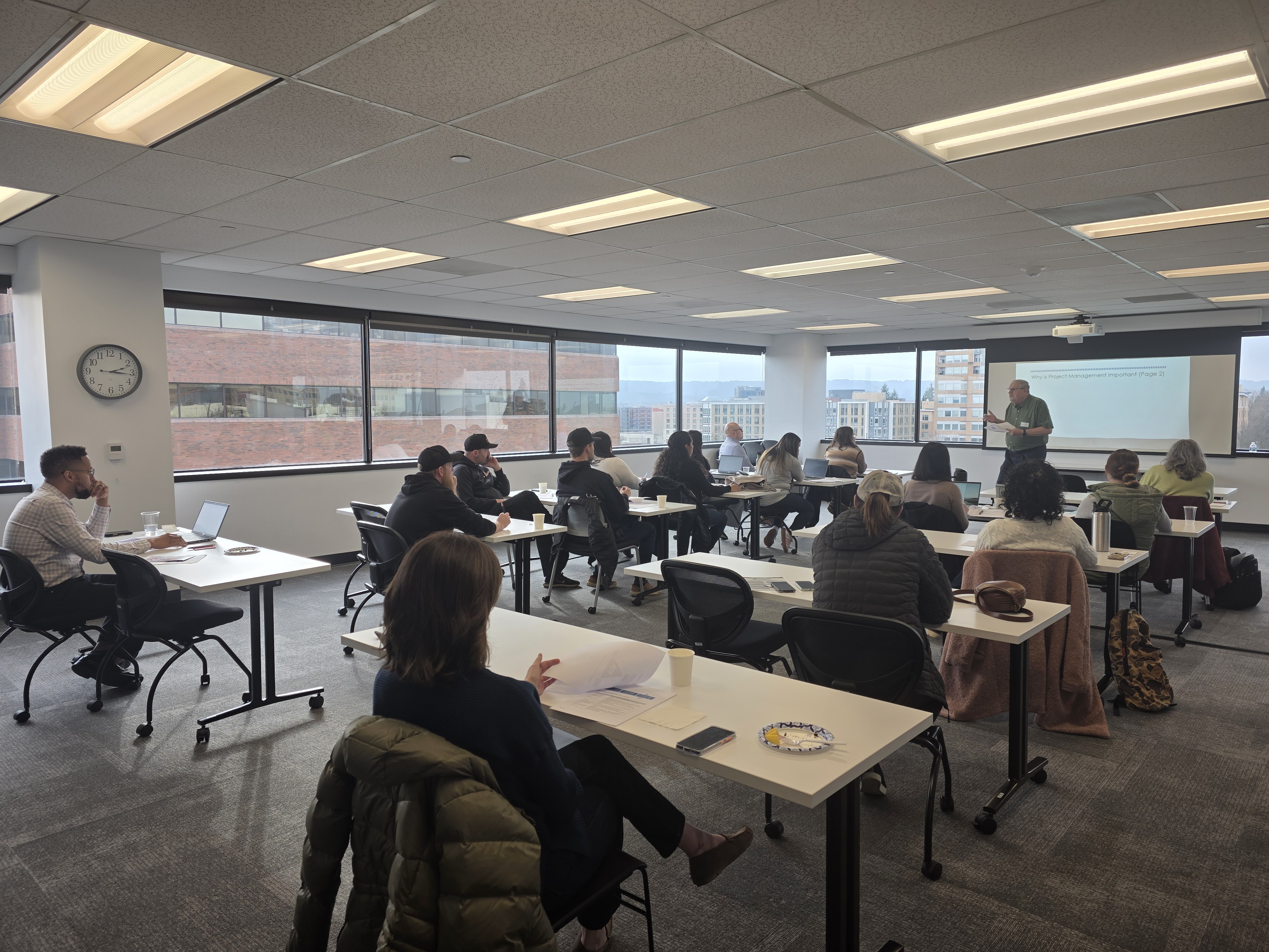 Dan Yeomans stands at the front of a room of business professionals as he presents a training on project management with a view of downtown Vancouver seen from the windows in the background