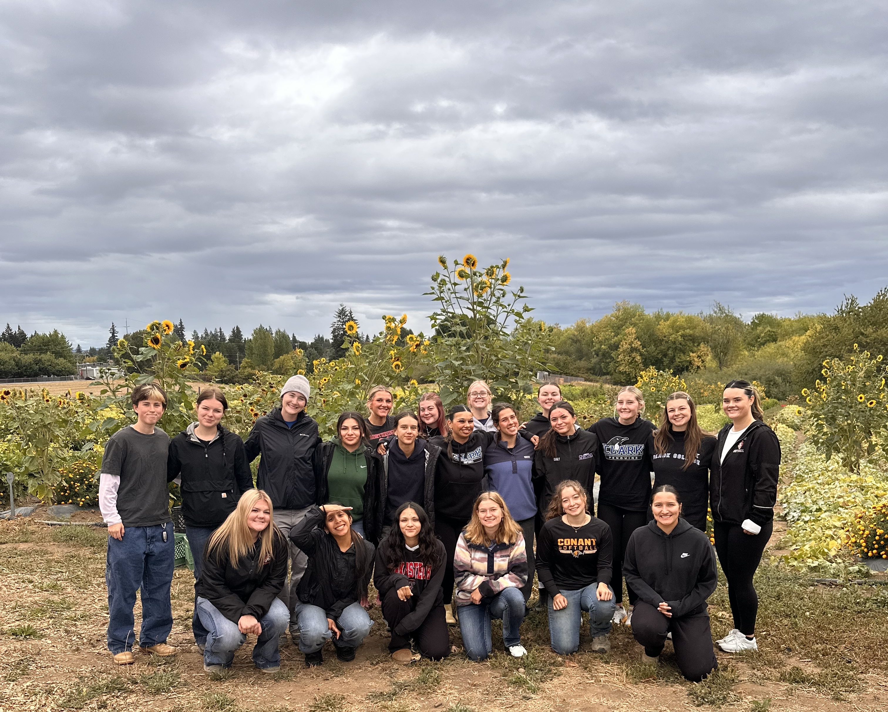 21 people pose together for a group photo. A farm is in the background, with tall sunflowers decorating the landscape.