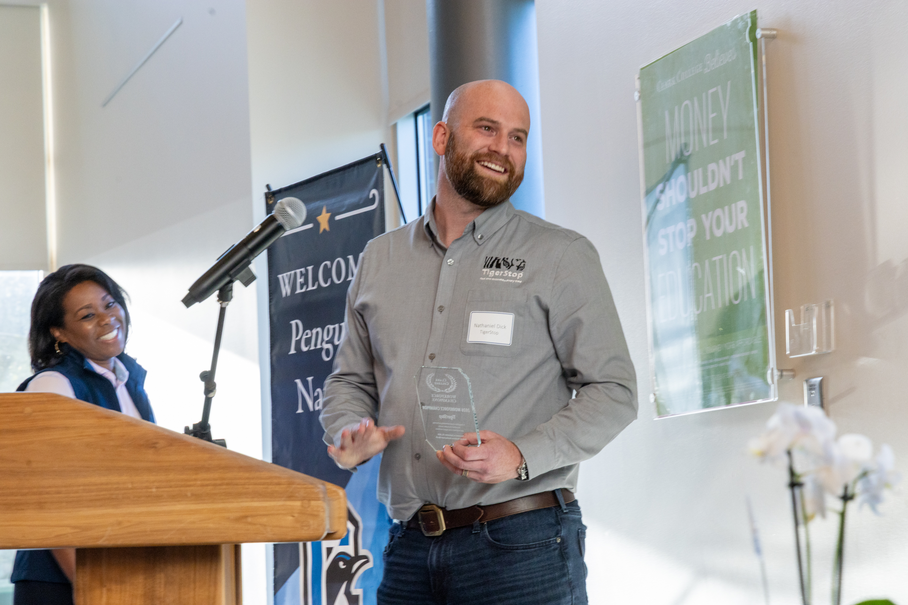 Nathaniel Dick smiles and holds the award at a podium. Melissa O'Dea smiles behind him.