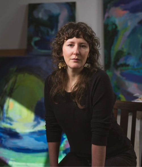 A person posing with a serious expression in front of a background filled with paintings displayed on the wall.