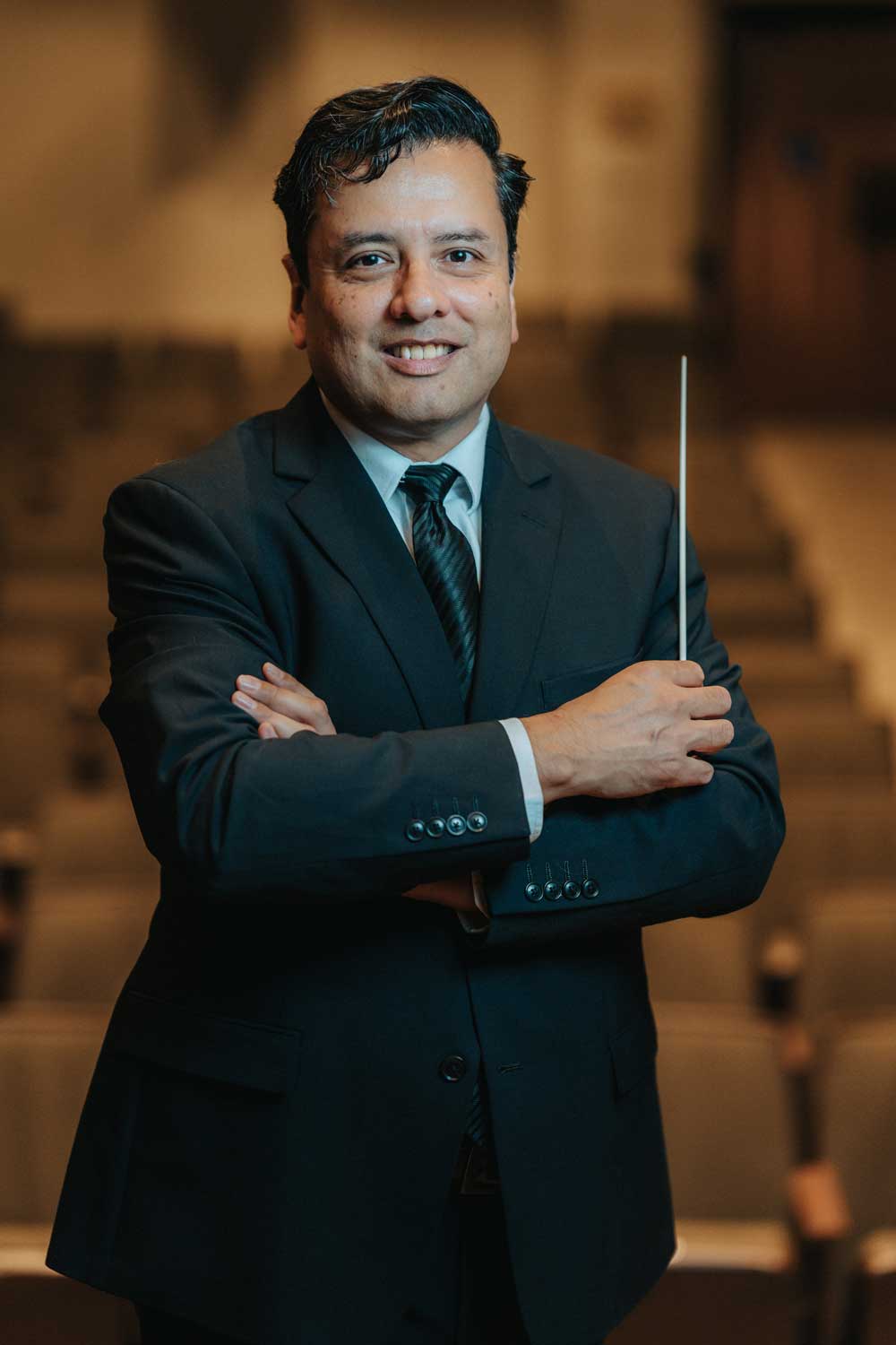 Portrait of an orchestra director (Dr. Rafael Antonio Rodriguez) in a suit, smiling at the camera while holding a conducting baton.