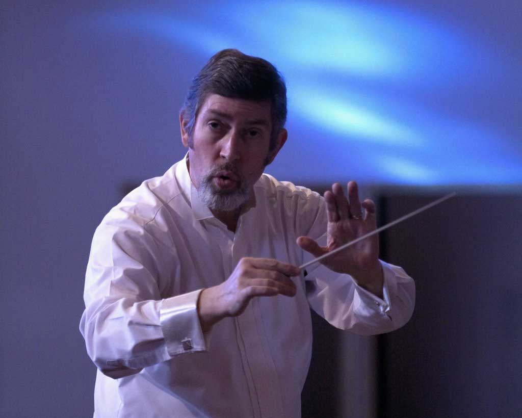 A conductor leading the orchestra ensemble, raising his baton to guide the performance.