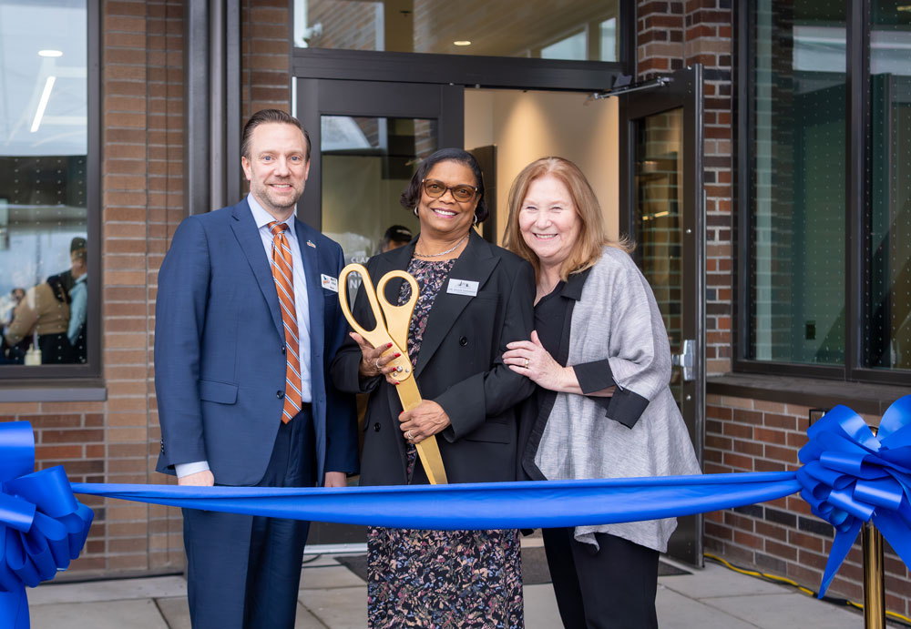 Three people stand in front of a building;  Dr. Edwards holds an oversized ceremonial key, with a ribbon stretched across two stands in front of them.
