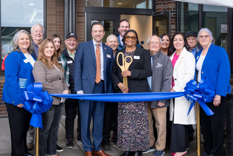 A group of people stands in front of a building; Dr. Edwards holds an oversized ceremonial key, and a ribbon stretches across two stands in front of them, ready to be cut.