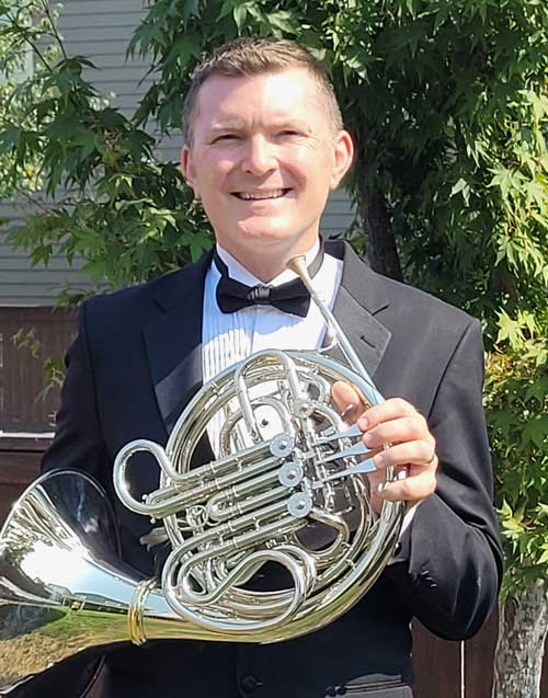 Photo of horn soloist, James Cameron wearing a suit and holding a french horn
