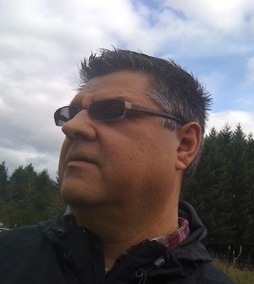 A side profile of a man wearing sunglasses, with a backdrop of tall pine trees