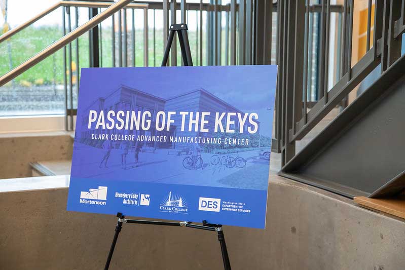 A poster board placed on an easel with the text 'Passing of the Keys Clark College Advanced Manufacturing Center' written on it. Below the carton, there is a row of sponsor names listed.