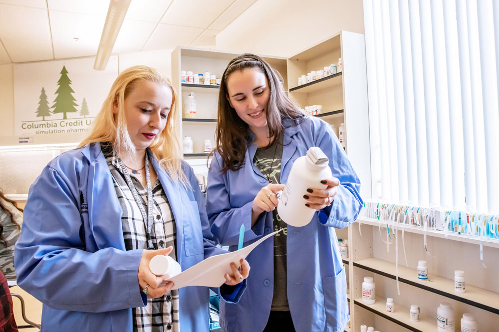 A pharmacy student examines a bottle closely while another student holds a sheet of paper. Together, they compare the bottle's label to the information on the page, with cabinets filled with supplies lining the wall behind them.