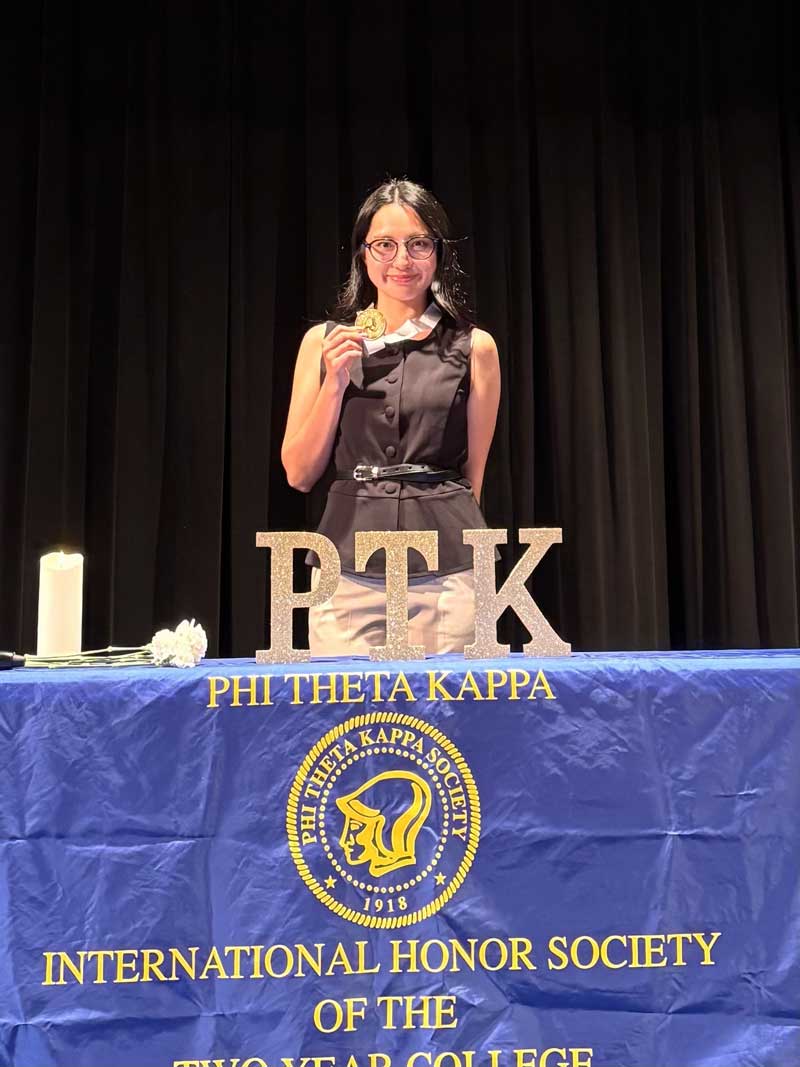A student wearing a circular gold medallion around their neck, standing behind a table displaying the letters 'PTK'