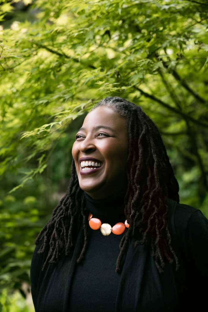 Author and poet Camille Dungy, smiling as she looks away from the camera in a forest setting.