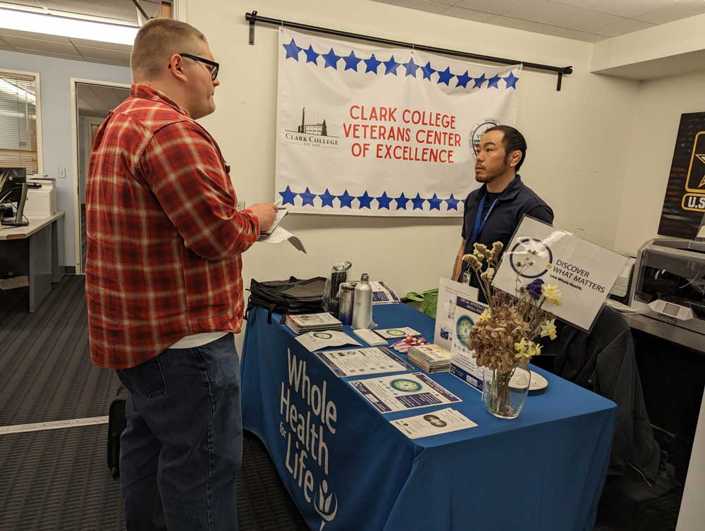 A person speaks with another person at a table covered with informational materials. A banner next to them reads 'Clark College Veterans Center of Excellence' and the tablecloth on the front of the table also displays the words 'Whole Health for Life'.